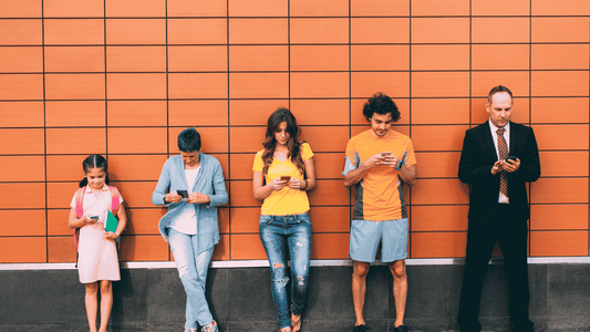 people of different ages looking down at their phones while leaning on a colorful wall.