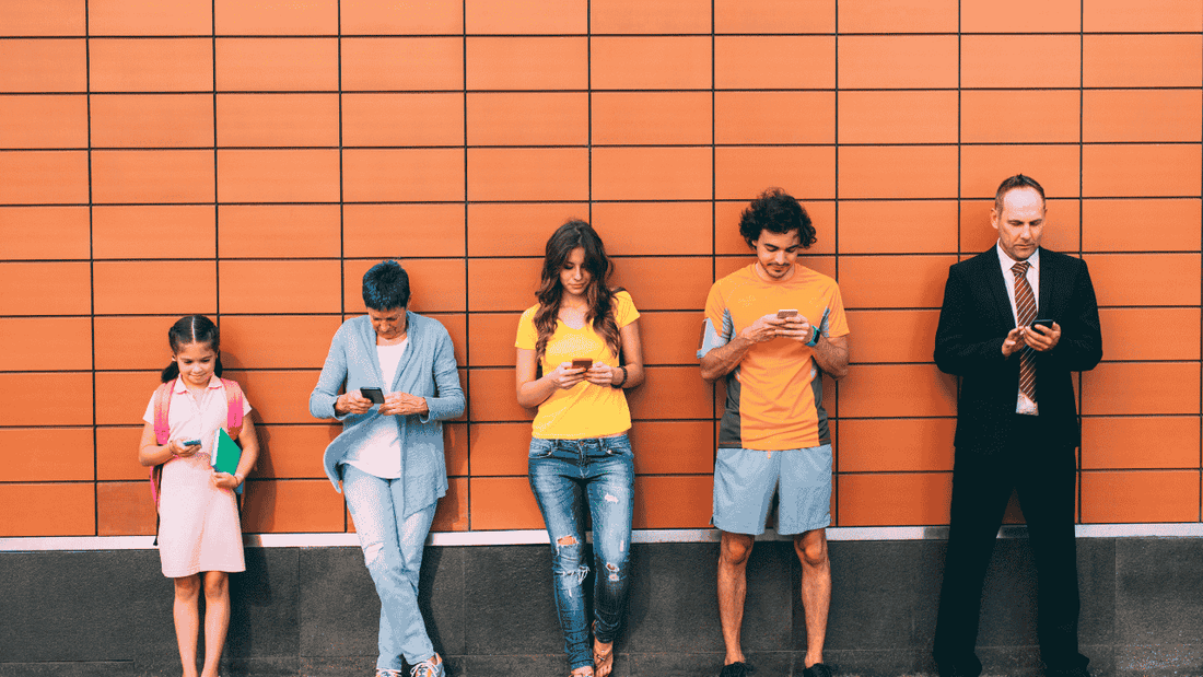 people of different ages looking down at their phones while leaning on a colorful wall.